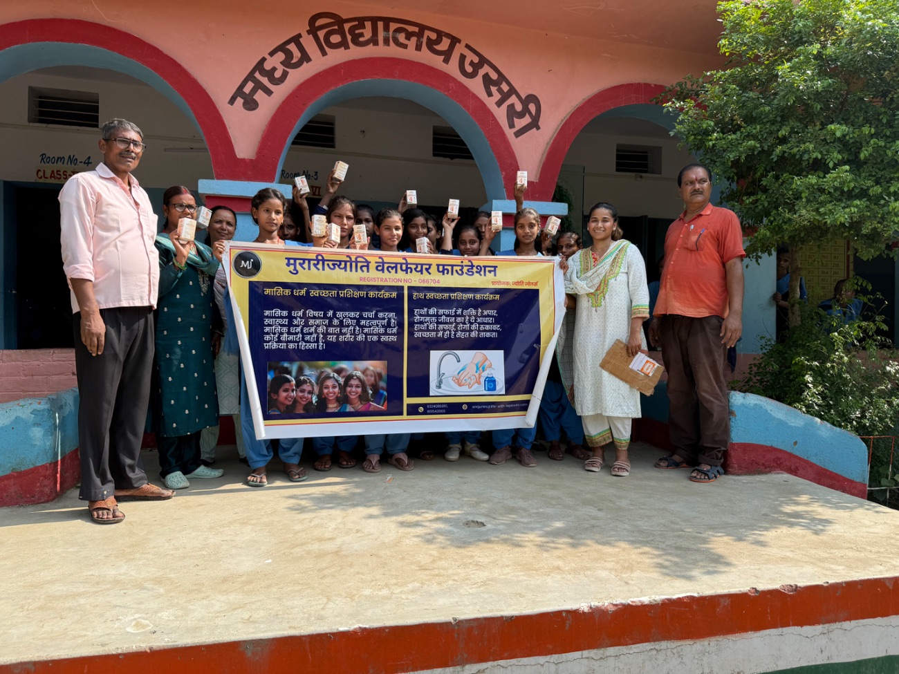 Schoolgirls hold up their menstrual cups outside their school beside an MJWF banner.