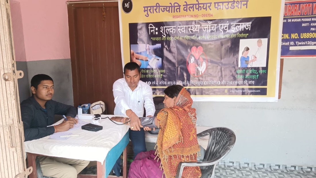 A woman has her blood pressure checked at a free MJWF health camp beneath the foundation banner.
