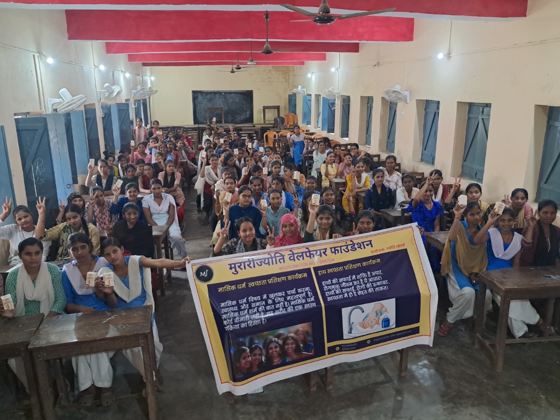 A school hall full of girls raising menstrual cups beside the MJWF awareness banner.